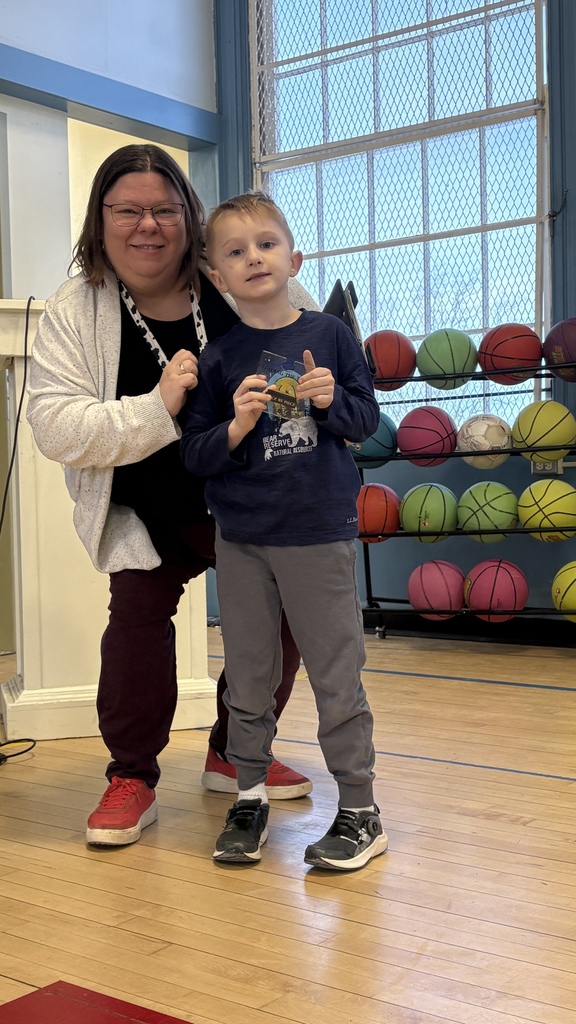 A young student stands beside a staff member while holding a small award, with a rack of colorful basketballs and gym windows behind them.
