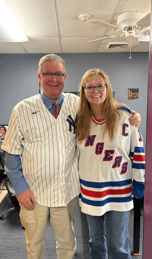 Two adults posing together indoors, wearing a Yankees baseball jersey and a Rangers hockey jersey.