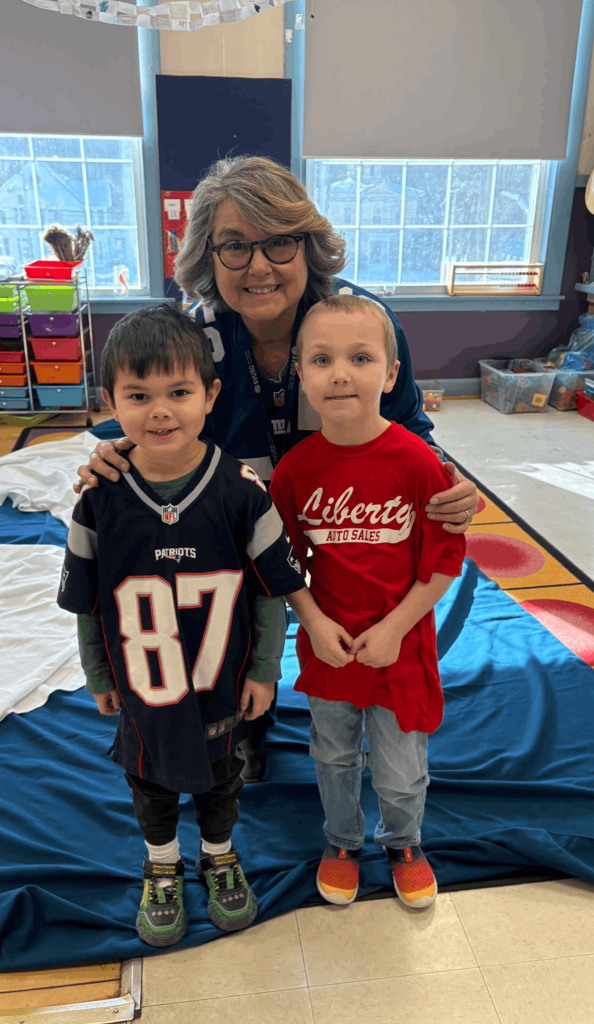 Teacher kneeling behind two young students wearing sports shirts, all smiling on a classroom rug.
