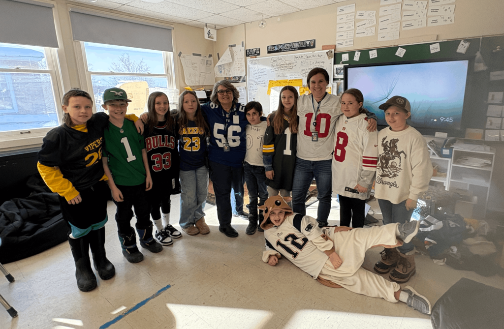 Group of students and two teachers in a classroom wearing sports jerseys, with one student playfully posing on the floor in front.