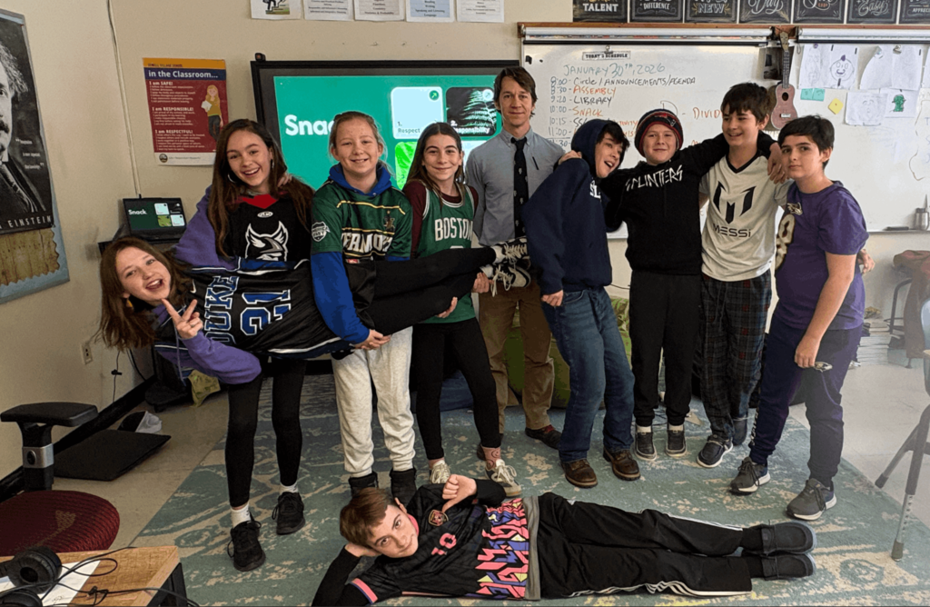 Group of middle school students in a classroom smiling while holding a classmate horizontally, with a teacher standing behind them near a whiteboard.