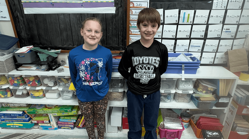Two elementary students standing side by side in front of shelves filled with math manipulatives and classroom supplies.