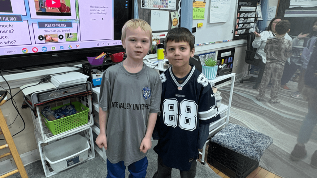 Two students standing in front of a classroom smartboard, wearing sports-themed shirts, with classmates visible in the background.