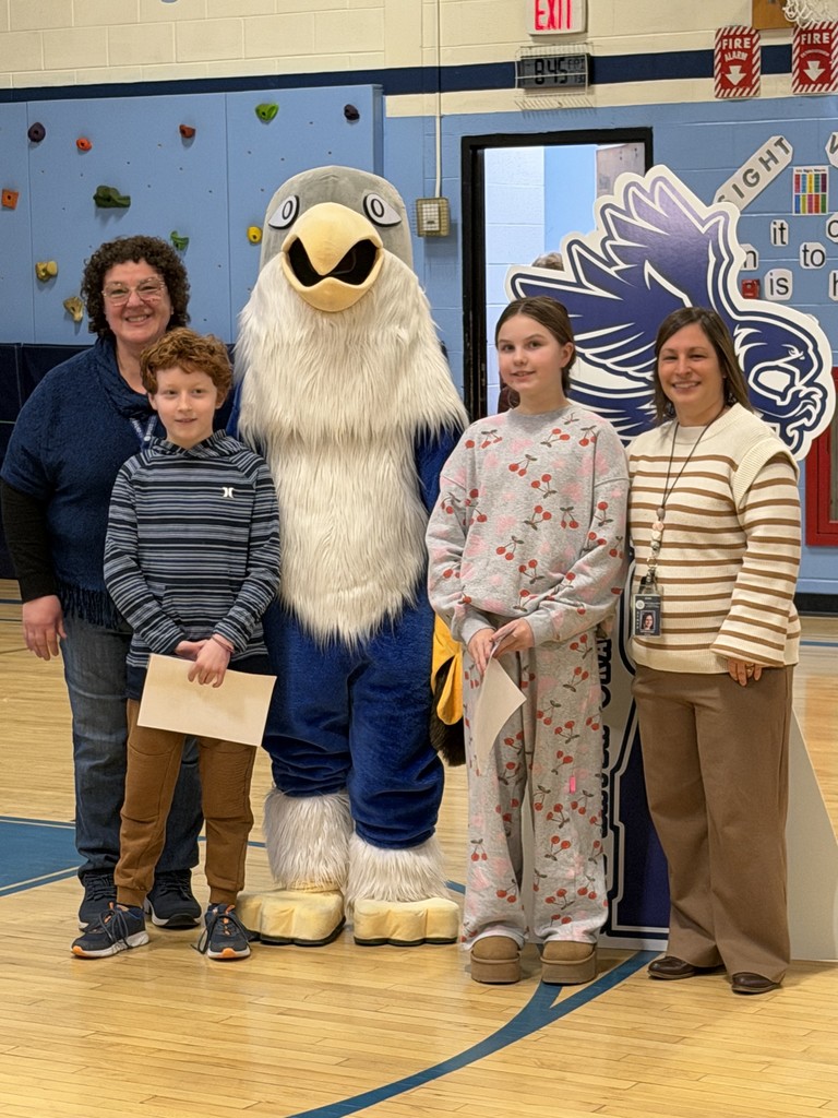 A group of five poses for a photo: two adult women, two children, and a blue-and-white falcon mascot. A young boy in a striped hoodie and a girl in a grey sweatsuit with cherry patterns stand in the center holding white papers. They are flanked by the mascot on the left and a woman in a striped cream sweater on the right.