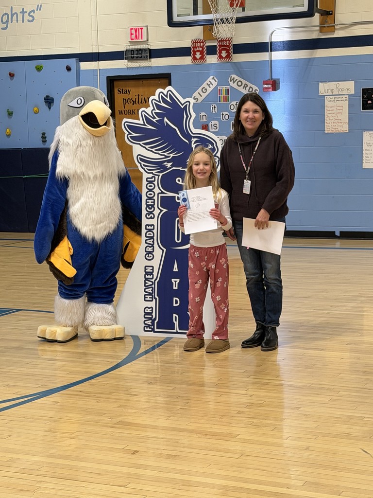 An adult woman and a young girl stand together in a gymnasium next to a "Fair Haven Grade School SOAR" falcon cutout. To their left, a blue-and-white falcon mascot stands with its wings slightly spread. Both the woman and the girl are holding white papers. The background features a basketball hoop, instructional posters, and a climbing wall.