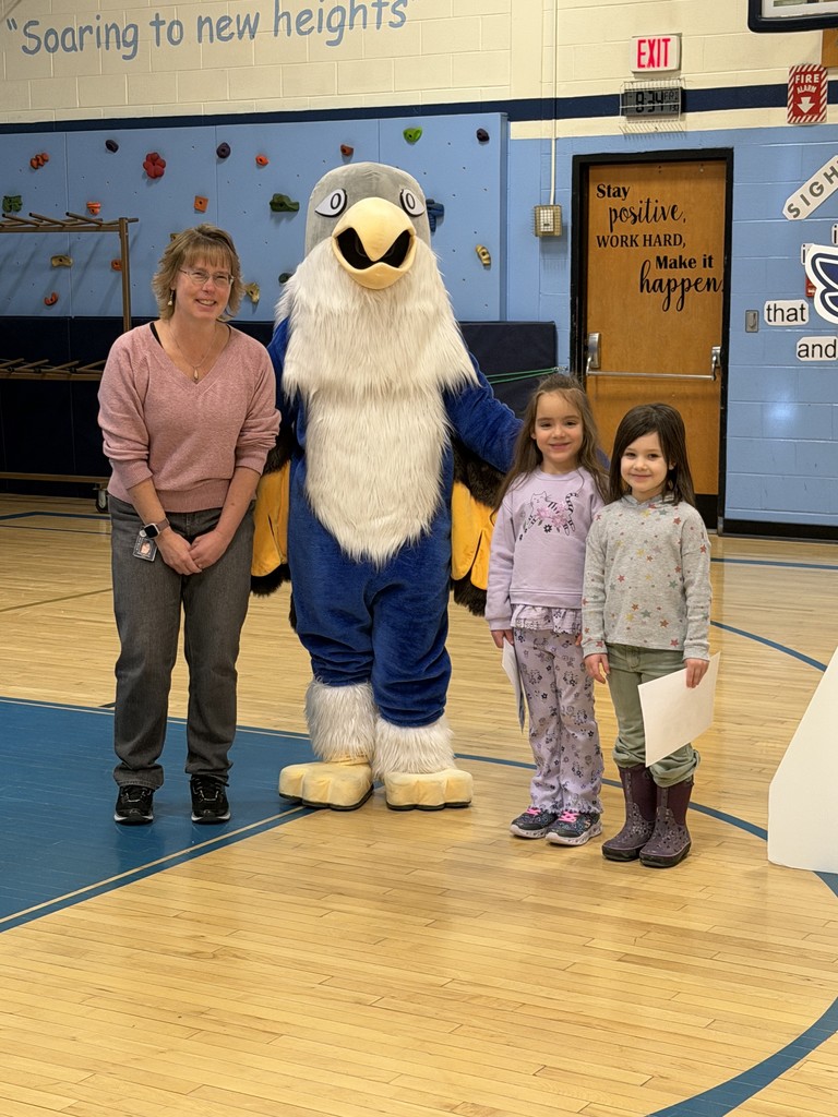 In a school gymnasium, an adult woman, a blue-and-white falcon mascot, and two young girls stand in a line on a wooden basketball court. The woman on the left wears a pink sweater and dark pants. The falcon mascot stands in the center with its wings slightly spread. To the right, two young girls stand side-by-side holding white papers. The background features a light blue wall with a small rock-climbing section and a wooden door with the words "Stay positive, WORK HARD, Make it happen."