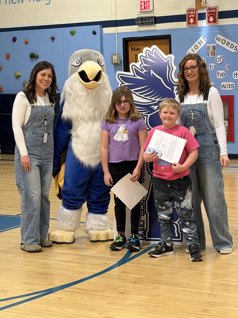 Two women in matching denim overalls and white long-sleeved shirts stand on either side of a blue-and-white falcon mascot and two children. The children, a girl in a purple shirt and a boy in a pink shirt, stand in front of a "SOAR" falcon cutout holding papers. The group is positioned on a wooden gymnasium floor.