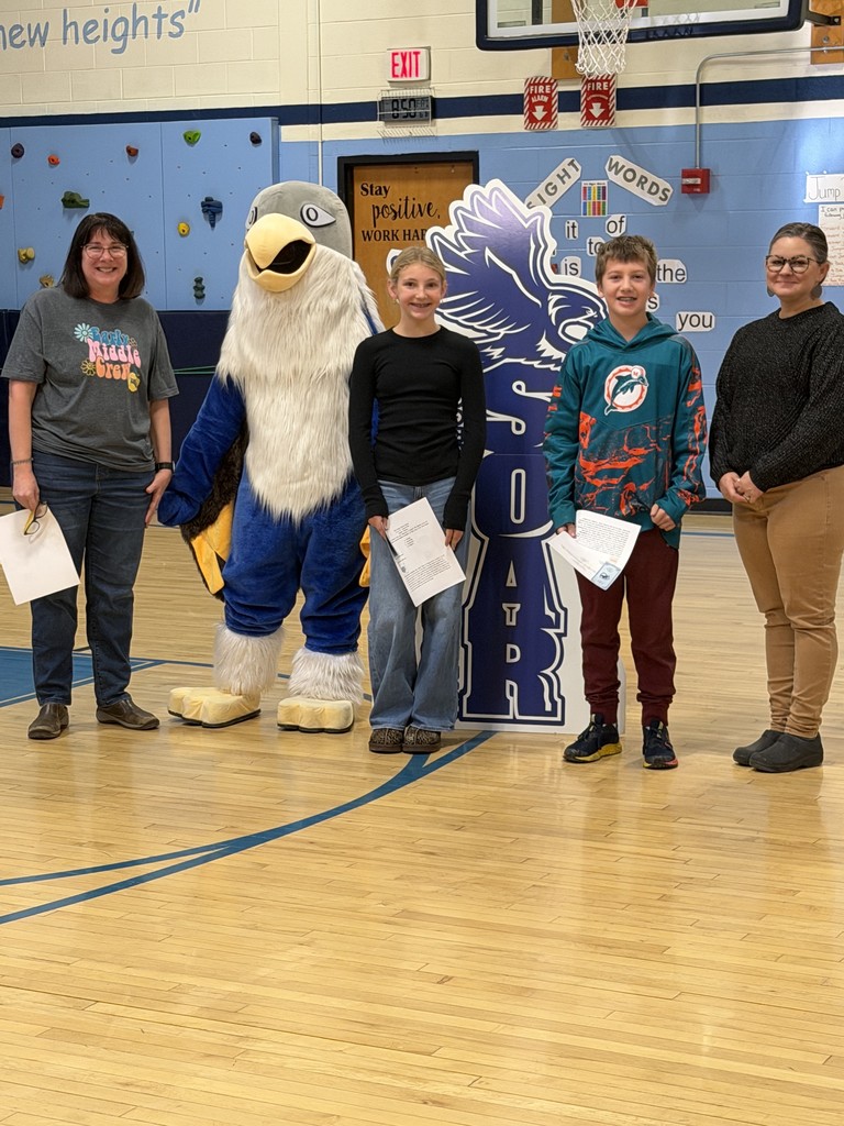Two women stand on the far sides of a group featuring a blue-and-white falcon mascot and two older children. The children, a girl in a black long-sleeved shirt and a boy in a teal hoodie, stand in front of the "SOAR" eagle cutout holding white papers. The woman on the left wears a grey t-shirt that says "Early Middle Crew".