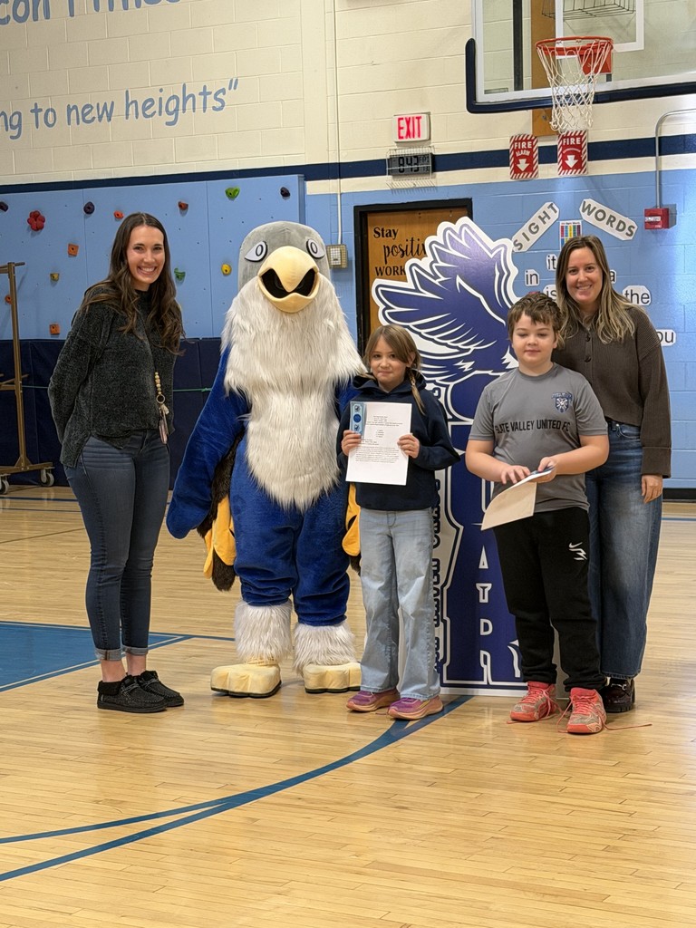 Two women stand on the outer edges of a group featuring a blue-and-white falcon mascot and two children. The children stand in front of a large "SOAR" falcon cutout, with the girl on the left holding a document. The group is positioned on a polished wooden gymnasium floor.