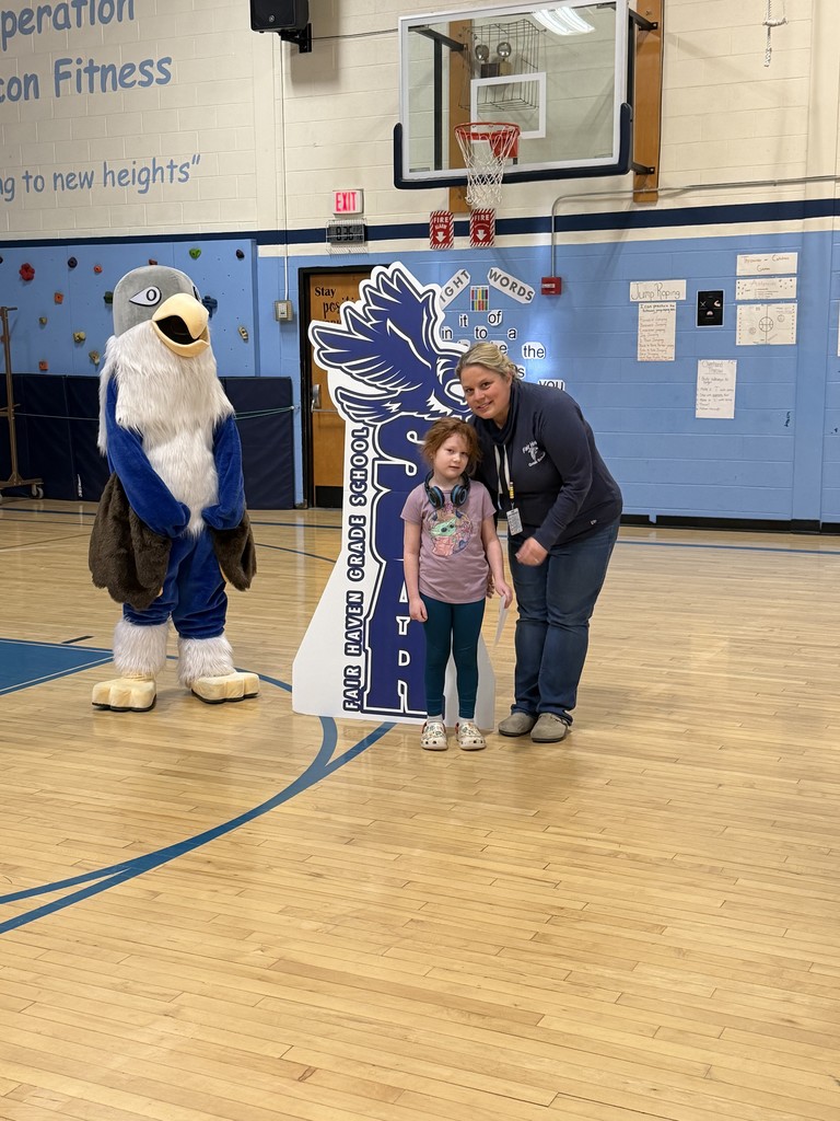 In a school gymnasium, an adult woman and a young girl stand next to a large cardboard cutout that reads "Fair Haven Grade School SOAR". To their left stands a blue-and-white falcon mascot with a white feathered chest. The girl wears a purple t-shirt, teal leggings, and headphones around her neck, while the woman wears a navy blue sweatshirt and jeans. The background features a basketball hoop, a climbing wall, and a blue wall with educational posters.