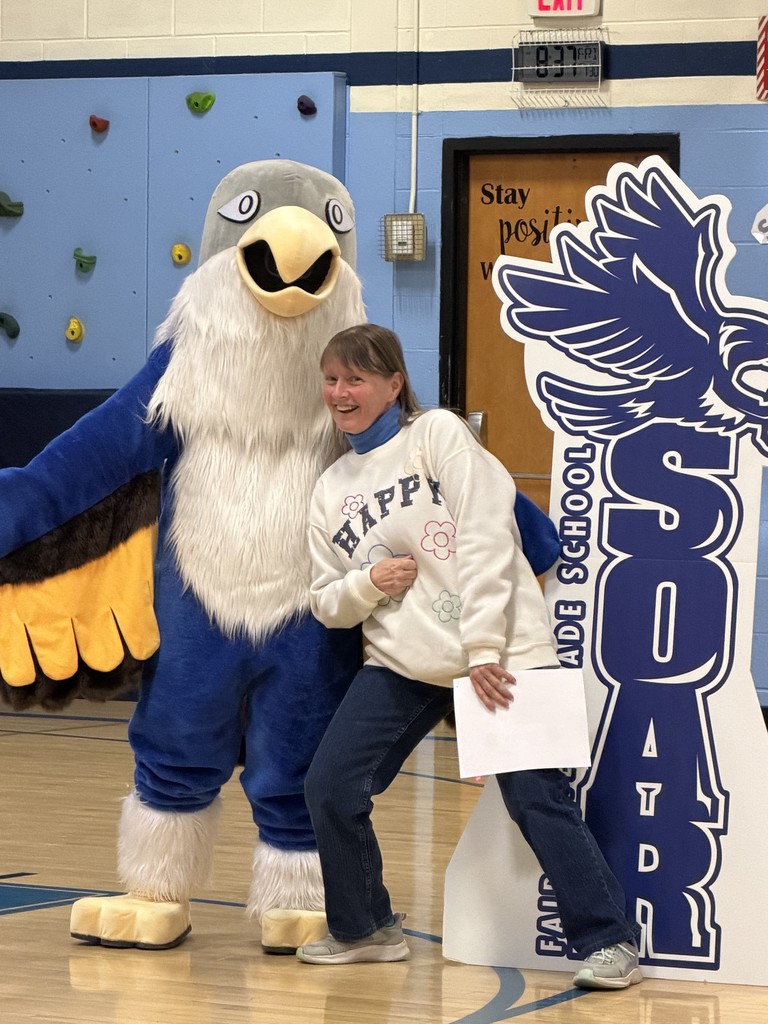 In a gymnasium, an adult woman in a white "HAPPY" sweatshirt and jeans poses playfully next to a blue-and-white falcon mascot. The woman is holding a white piece of paper and is positioned between the mascot and a large white-and-blue "SOAR" cutout. Behind them is a blue wall featuring a climbing section with colorful grips and a wooden door with motivational text.