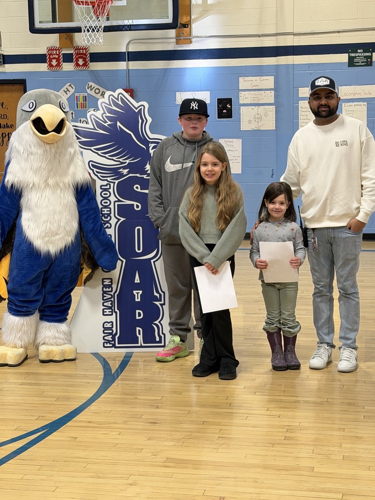 A mascot in a blue and white falcon costume stands next to a large "SOAR" cutout. Three students and a man wearing a baseball cap and a white sweatshirt pose for a photo on a gymnasium floor.
