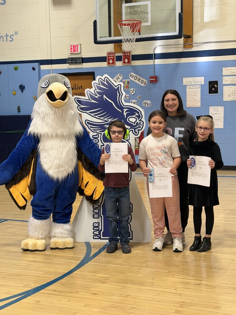A school mascot and a female faculty member stand with three elementary-aged students. The students are holding certificates and small blue ribbons while posing in front of a blue wall featuring "Sight Words.
