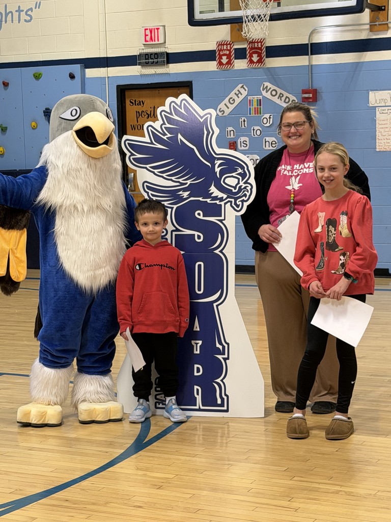 A mascot in an falcon costume, a woman in a pink shirt, and two children pose together. The boy in a red hoodie and the girl in a red patterned sweater are holding award papers in a school gym.