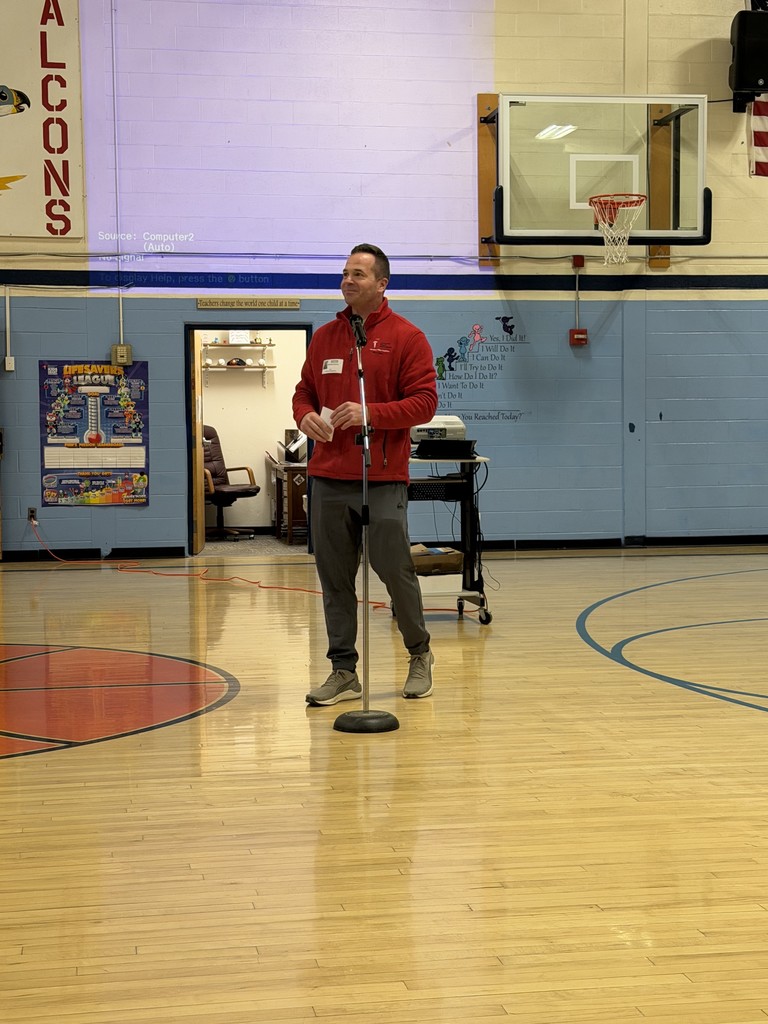 A man in a red zip-up jacket and grey athletic pants stands at a microphone in the center of a gymnasium. He appears to be speaking to an audience during a school assembly or event.