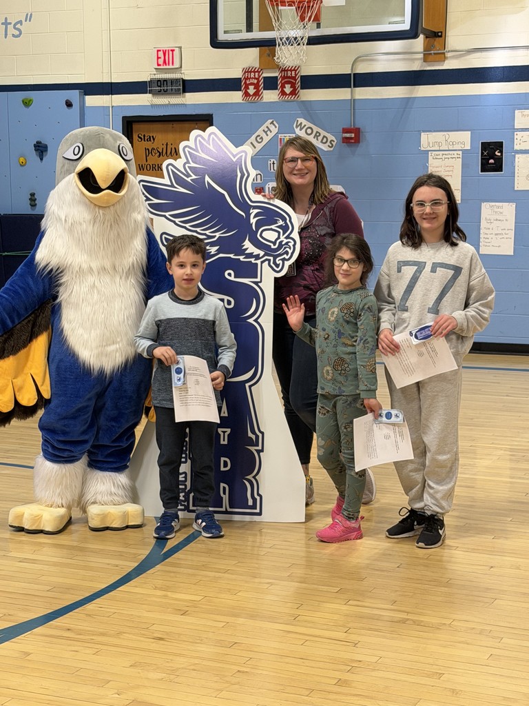 A group photo featuring the falcon mascot, a female teacher in a purple shirt, and three students. One student wears a grey sweatshirt with the number "77" on it; they are all holding award certificates.