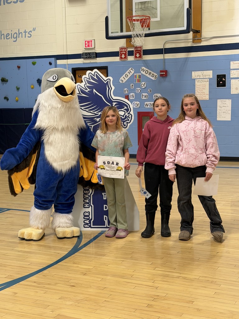 The falcon mascot poses with three young girls in a gymnasium. One girl holds a large "SOAR" certificate featuring school bus illustrations, while the others hold smaller award papers.