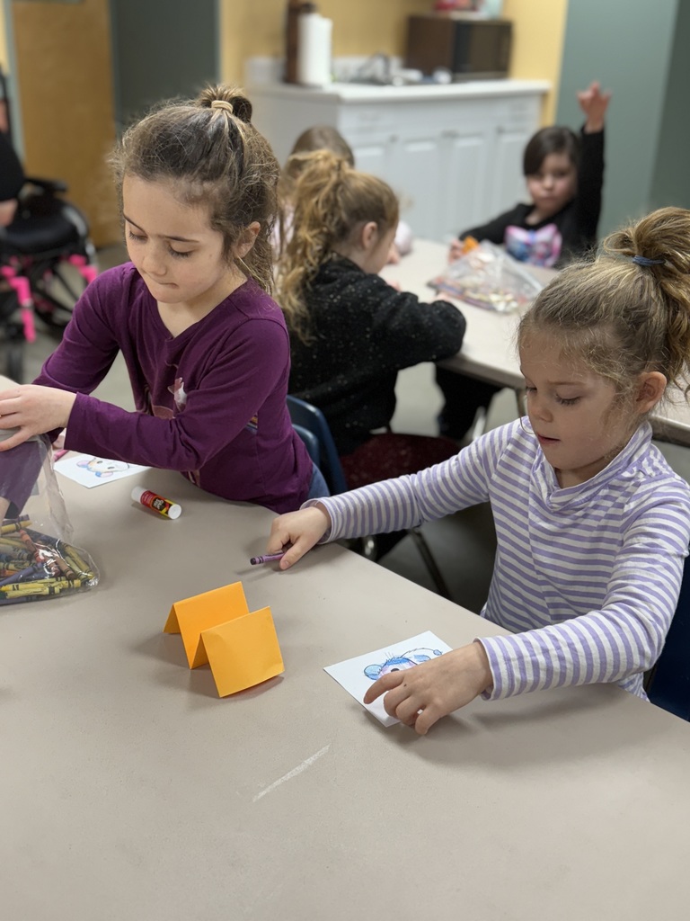 Students sit around a table coloring while another child raises a hand in the background.