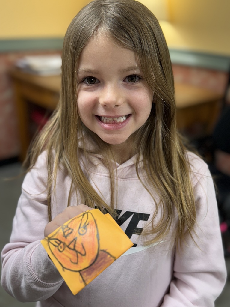 Student smiles while showing a colored paper craft with a hand-drawn face design.