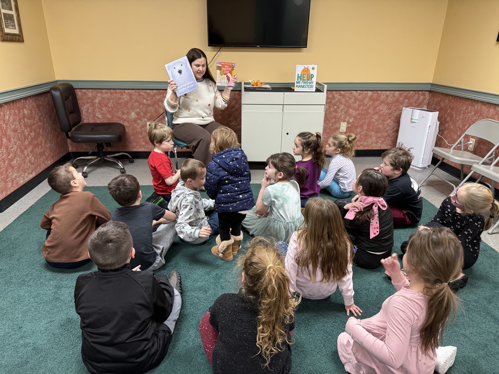 Adult librarian reads aloud to a seated group of elementary students gathered on a carpet for story time.