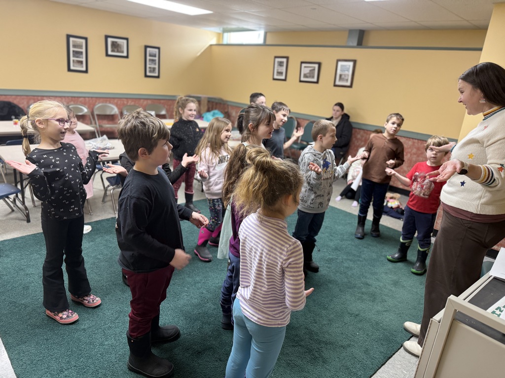 First grade students stand in a circle with a librarian, participating in a movement and song activity.
