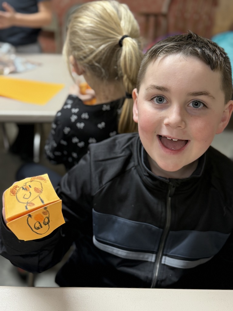 Smiling student holds up a folded paper craft decorated with a drawn face.