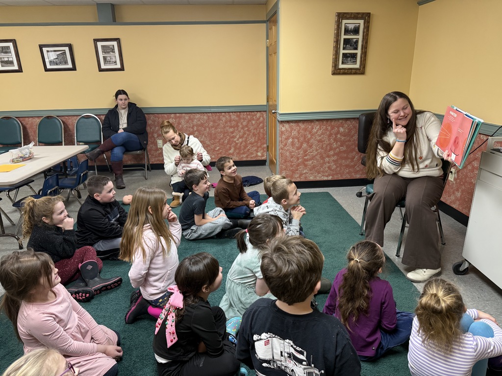 Children sit on the floor listening and smiling as a librarian reads from a picture book at the front of the room.