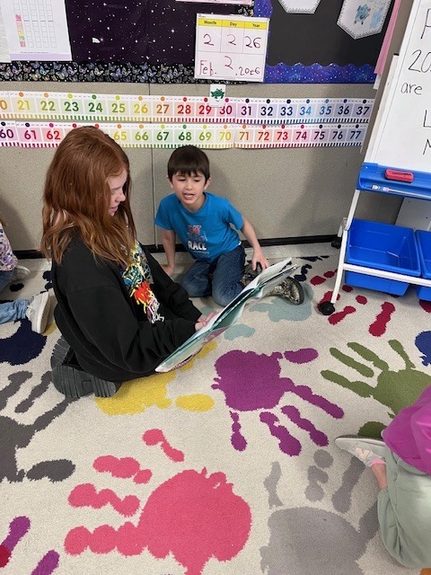 Two students sit on a colorful rug featuring large, multi-colored handprints. An older student with long red hair holds a large picture book open while a younger boy in a blue t-shirt watches and listens. A classroom calendar and number line are visible on the wall behind them. 