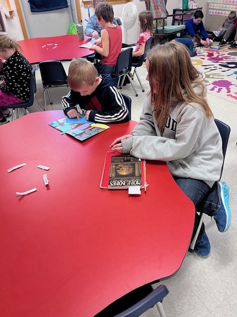  A wide shot of a classroom showing students working at red tables and on the floor. In the foreground, a boy in a navy hoodie reads a picture book while a girl in a grey hoodie sits next to him with a book titled "More Bones" on the table. 