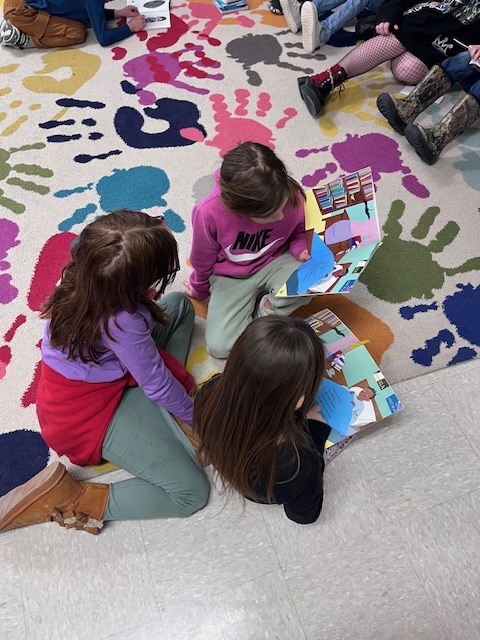  A high-angle shot of three girls sitting in a close circle on the handprint rug. Each girl is holding or looking at colorful picture books, appearing to read together or share stories in a small group. 