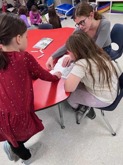  Three students are gathered around a bright red, kidney-shaped table. An older student wearing glasses points to a page in a book, guiding two younger girls who are focused on the text. Small slips of paper are scattered on the table.
