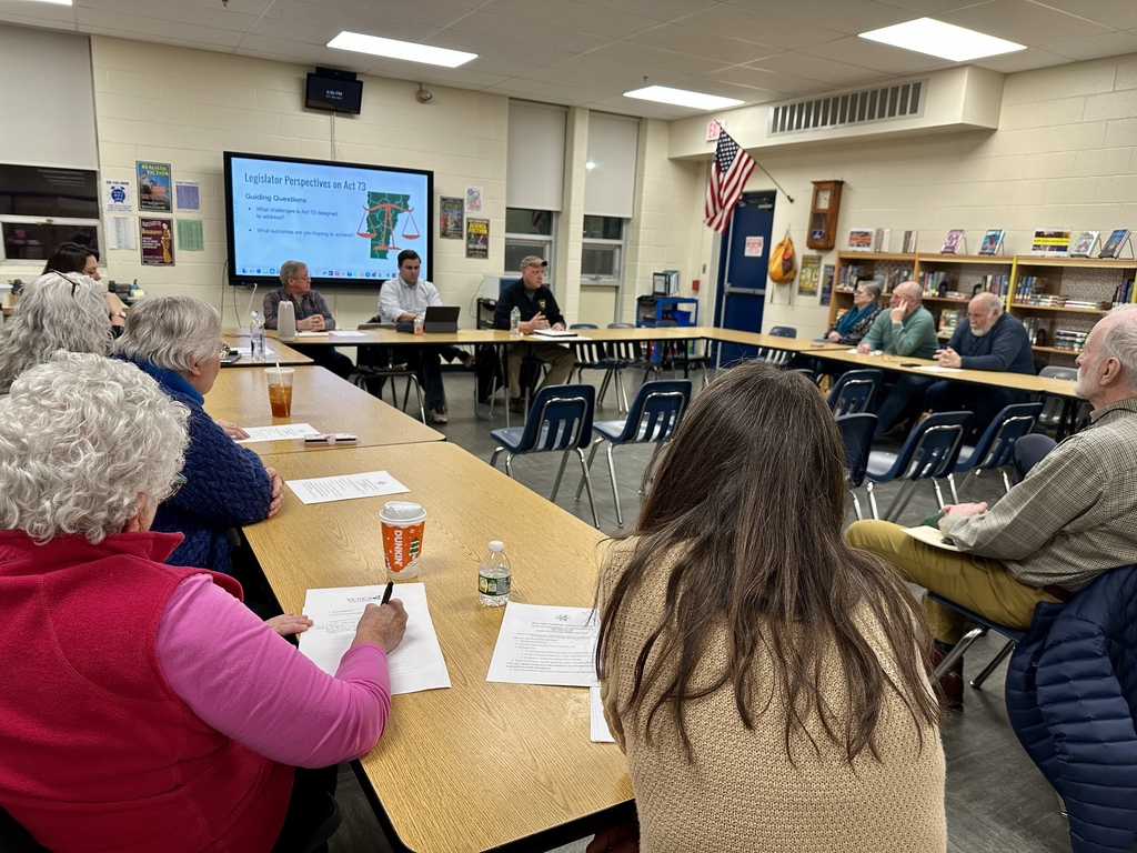 Community members and local legislators sit around tables in a school library classroom during a public discussion meeting, with a presentation on a screen about Act 73 and participants taking notes.