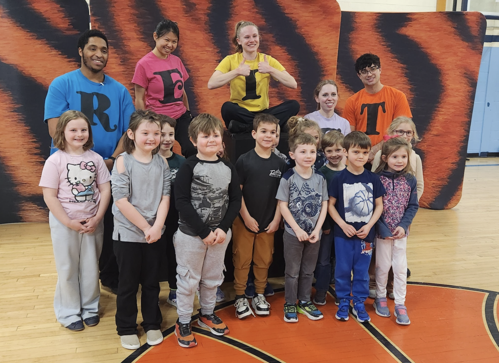 Group of elementary students and several adult presenters posing in a school gym in front of an orange-and-black backdrop, smiling for a photo; the adults wear bright shirts with large letters that spell a word together.
