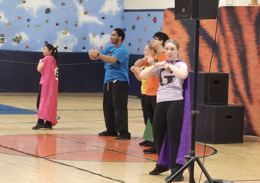 Four presenters in colorful shirts and capes performing coordinated motions at the front of a school gym during a student assembly.