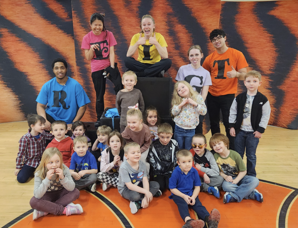 Larger group of young students and adult presenters gathered in a gym, smiling and posing together in front of the same orange-and-black backdrop, with some presenters giving thumbs-up.