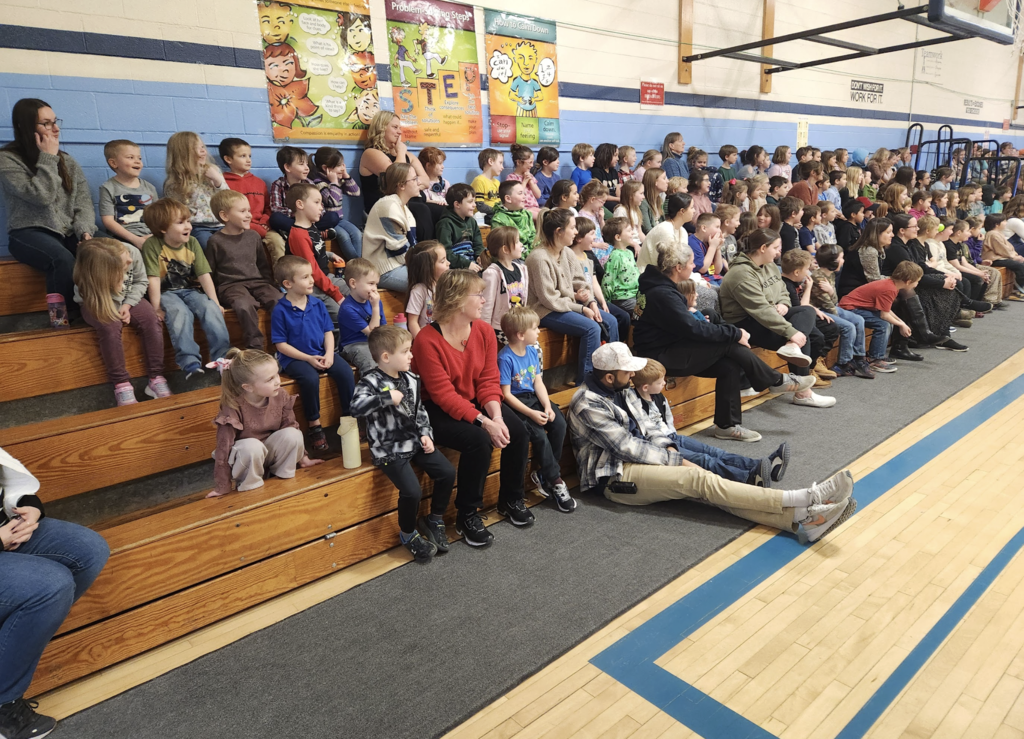 Dozens of elementary students and several adults seated on wooden gym bleachers, watching an assembly or presentation off-camera.