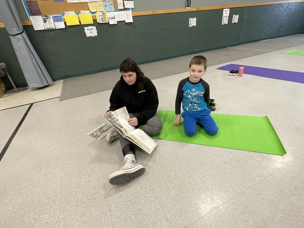 A teenage girl with long dark hair sits on a gray rug next to a bright green mat, holding open a large picture book. A young boy in a blue Transformer shirt sits on his knees on the mat next to her, looking toward the camera. 