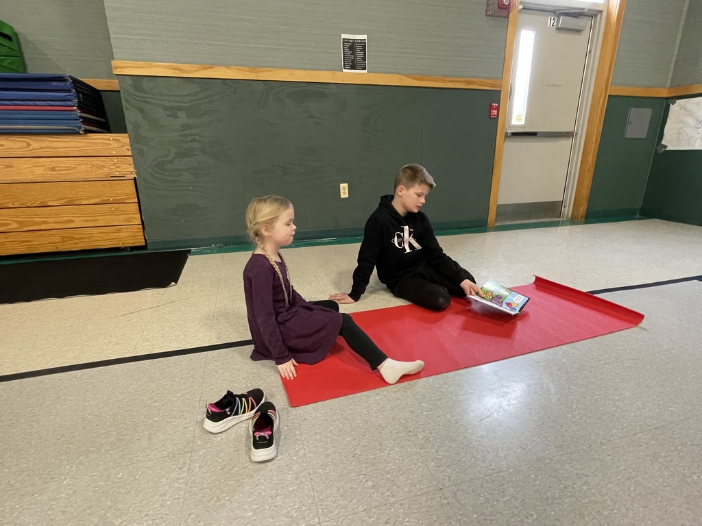  A young girl with blonde braids sits on a red mat, looking toward a teenage boy in a black "Calvin Klein" hoodie. The boy is leaning back and holding a colorful book open for her to see. Her shoes are placed on the tile floor next to the mat.  