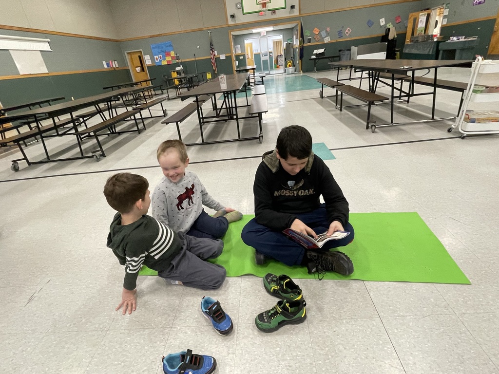  An older boy wearing a black "Mossy Oak" hoodie sits cross-legged on a bright green mat in a school cafeteria, reading a book to two younger boys. The younger boys sit closely, looking at the pages with smiles. Several pairs of shoes are scattered on the floor in front of them.  