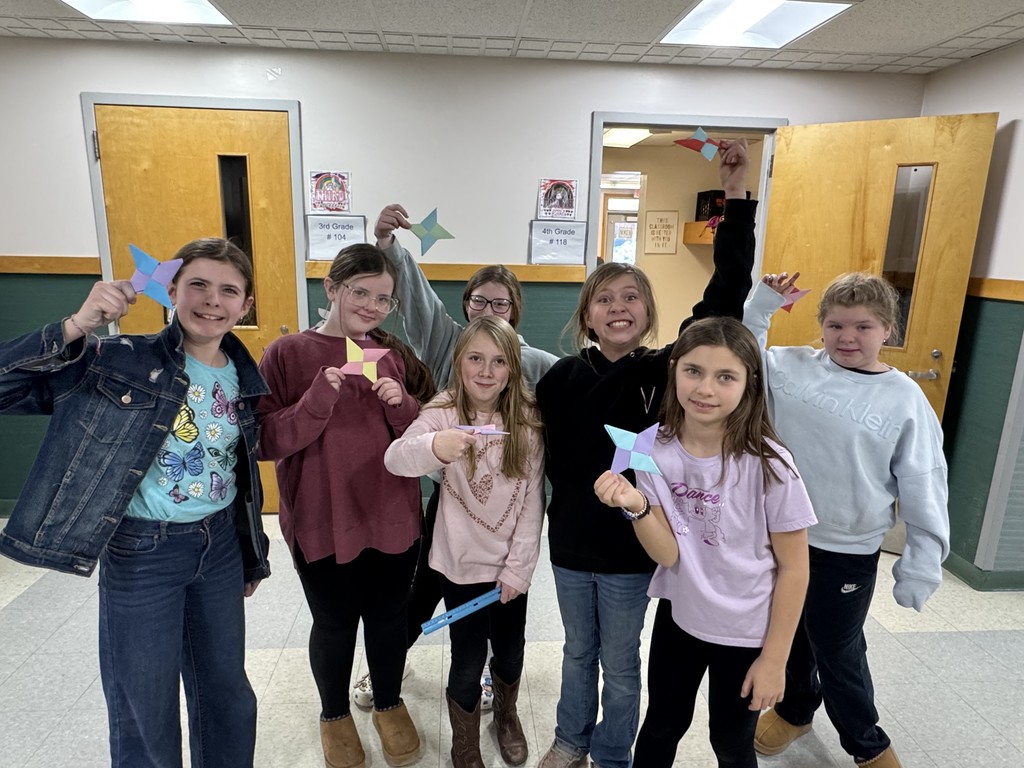  Seven smiling girls stand together in a school hallway, proudly displaying their completed multi-colored origami ninja stars. They are posed in front of two classroom doors labeled "3rd Grade" and "4th Grade." The group shows a range of expressions from broad grins to quiet smiles, with some holding their paper creations high in the air.