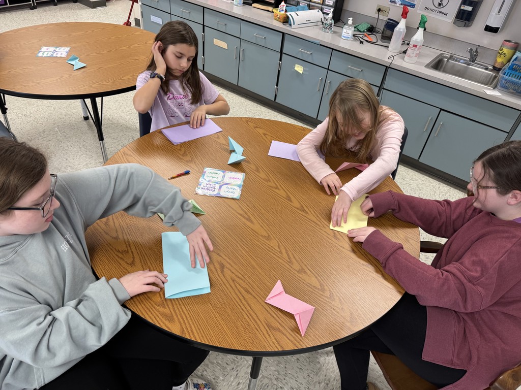 Four young girls sit around a circular wooden table in a classroom, focused on folding colorful sheets of paper. They are in various stages of an origami project, with light blue, purple, yellow, and pink paper visible. One girl in a grey hoodie carefully creases a large blue sheet, while her peers work on smaller sections and pre-folded shapes.  