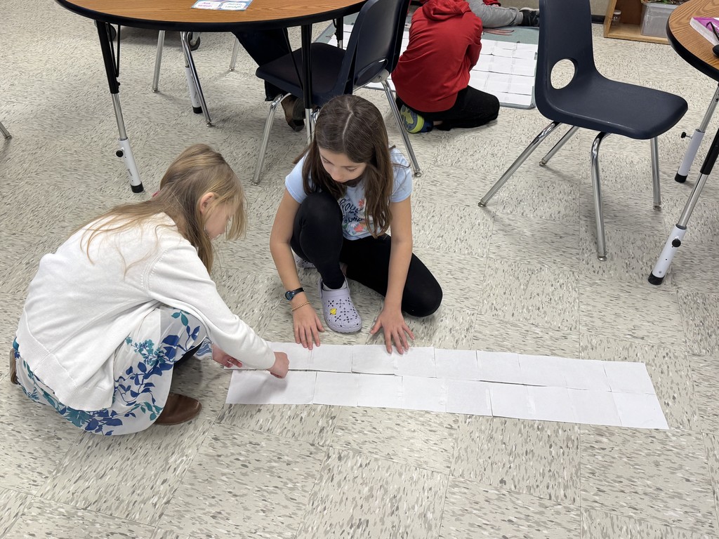Two young girls kneel on a classroom floor, working together to align and tape two long horizontal rows of white grid paper. They are focused on making sure the edges of the squares meet perfectly.  IMG_1740.jpg Two boys in red hoodies sit on a colorful rug featuring a map of the United States. They are assembling several long strips of grid paper side-by-side to create a large rectangular mat on the floor. 