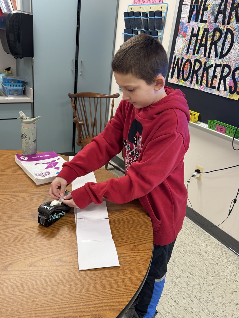 A young boy in a red hoodie stands at a wooden classroom table, carefully using a black tape dispenser to join together squares of white grid paper into a long strip. A purple "Bridges in Mathematics" student book lies on the table nearby.  