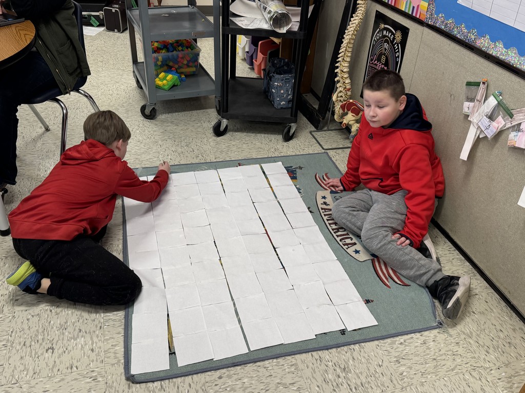 Two boys in red hoodies sit on a colorful rug featuring a map of the United States. They are assembling several long strips of grid paper side-by-side to create a large rectangular mat on the floor.  