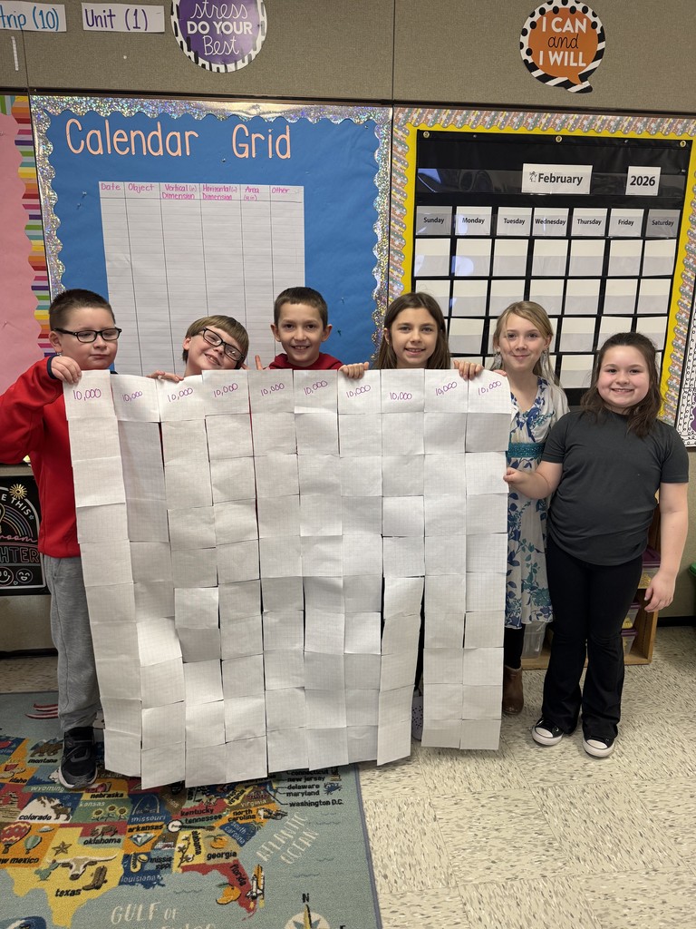  Six smiling students stand in a line in their classroom, proudly holding up a large, completed grid made of taped-together paper squares. Each column of the grid is labeled "10,000" at the top in purple marker. In the background, a "Calendar Grid" bulletin board and a February 2026 calendar are visible.