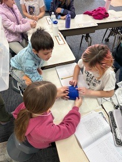 Four elementary students gathered around desks working together with small blue containers and tools during a hands-on classroom activity.