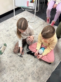 Two elementary students sit on a classroom rug using a small hammer and tool to test or assemble a simple object while classmates sit nearby observing.