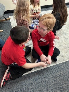 Two young students sitting on the floor facing each other and talking while working with a small object during a partner activity.