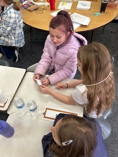 Three elementary students at a desk examining small white objects and containers while recording notes on clipboards.