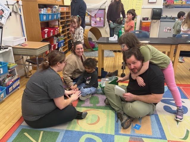 Several adults and young students gathered on a classroom rug working together on a hands-on activity, with shelves of supplies and play materials in the background.